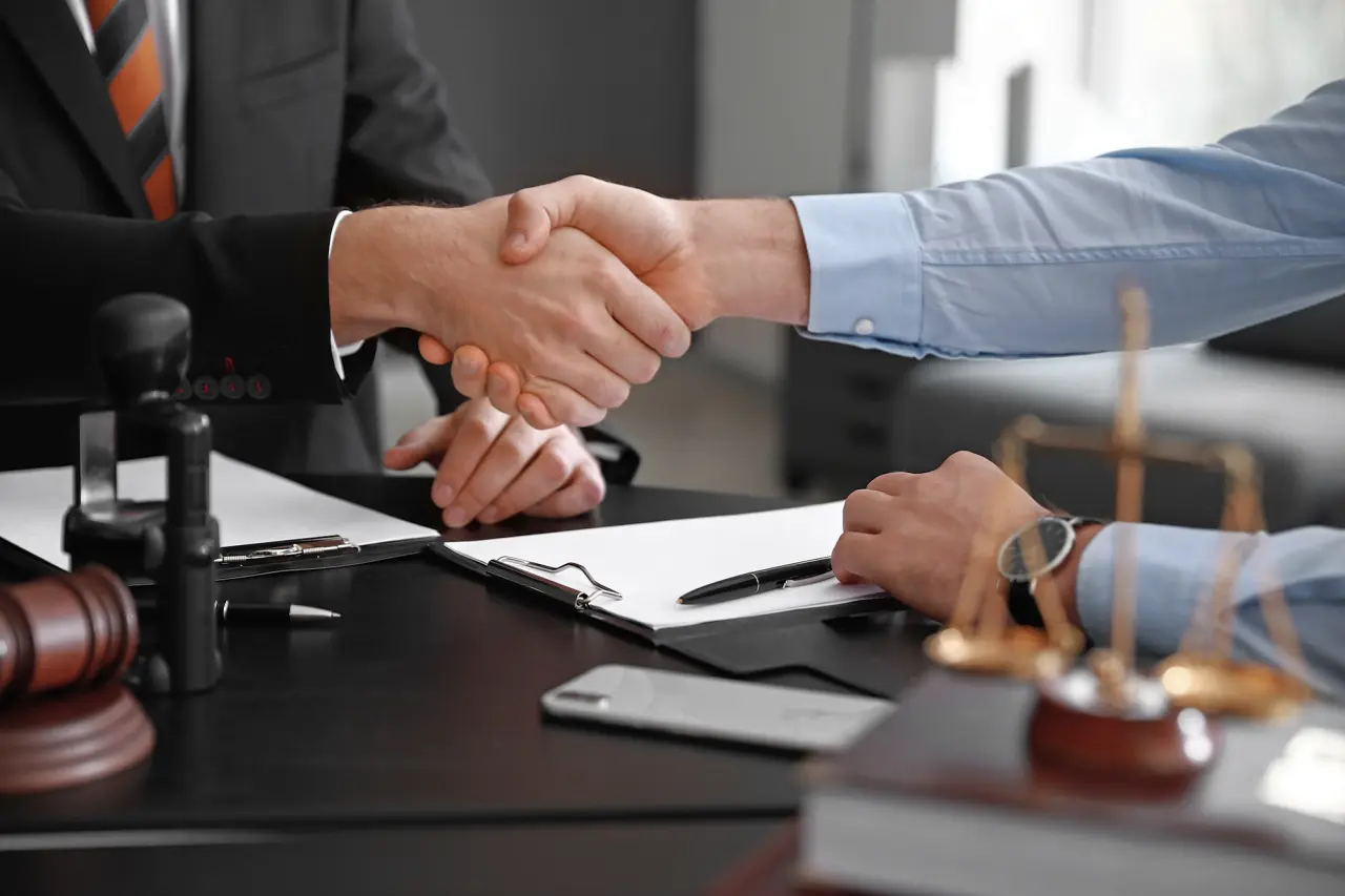 Two people shaking hands over a table with paperwork on it